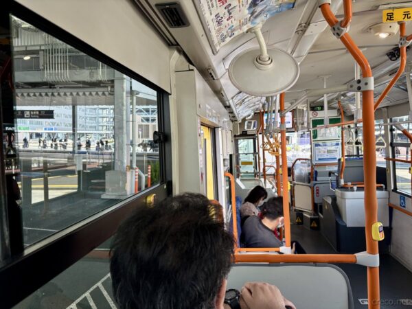 成田空港行きの連接バス車内から望む大型ターミナルビルのガラス壁。空港へ向かう乗客が座り、窓の外に到着ロータリーが見える。沖縄旅行の出発シーン。