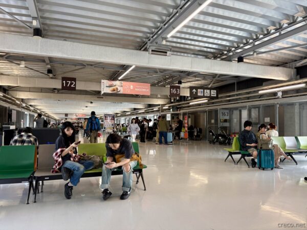 成田空港第2ターミナル出発ロビー172番と163番付近のベンチで搭乗を待つ旅行者たち。吹き抜けの高い天井と案内表示、搭乗前の空港風景。