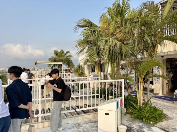 瀬長島ウミカジテラスから滑走路方向を望む展望デッキ。ヤシの木越しに那覇空港が見え、旅行者がカメラを構える夕暮れのひととき。