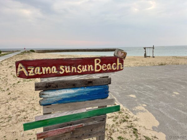 あざまサンサンビーチの木製看板「Azama sunsun Beach」。流木調の支柱に赤いペイント文字、朝焼けのビーチ入口の目印。