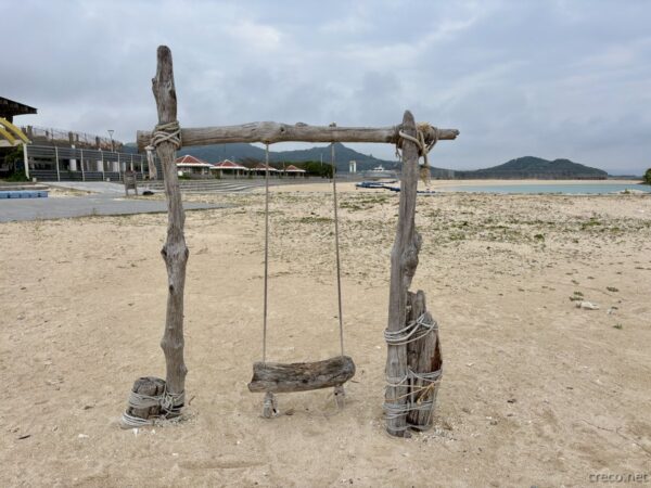 あざまサンサンビーチのブランコ越しに広がる砂浜。奥に玉城の集落と山並みが見え、南城市の朝のビーチ全景。