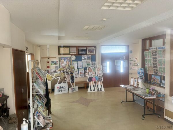 具志川公民館ロビー内観。沖ツラのキャラクター等身大パネル（比嘉律と喜屋武飛夏）が展示され、観光パンフレットが並ぶ聖地巡礼の拠点。