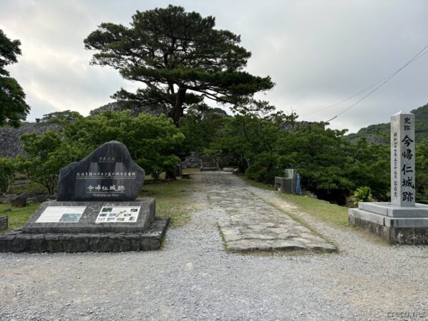 世界遺産・今帰仁城跡の入口広場。石碑と案内板、奥に木々と城壁、琉球王国時代の三山の一つ北山の居城跡。