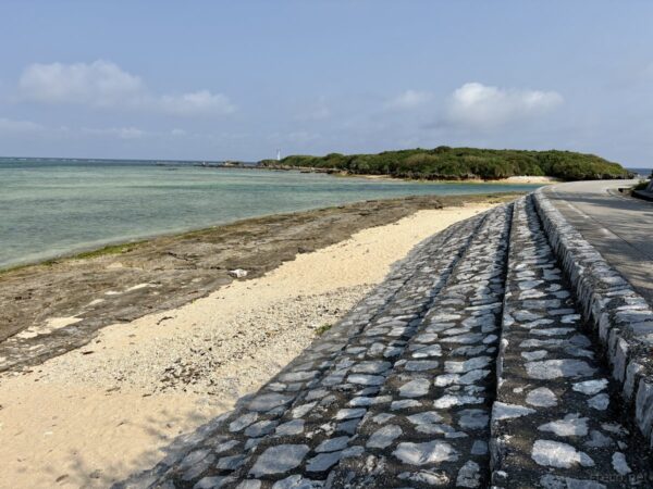 備瀬崎の石畳の護岸と遊歩道。海辺の石垣と白砂ビーチ、奥に灯台、本部町の最北端の海岸ポイント。