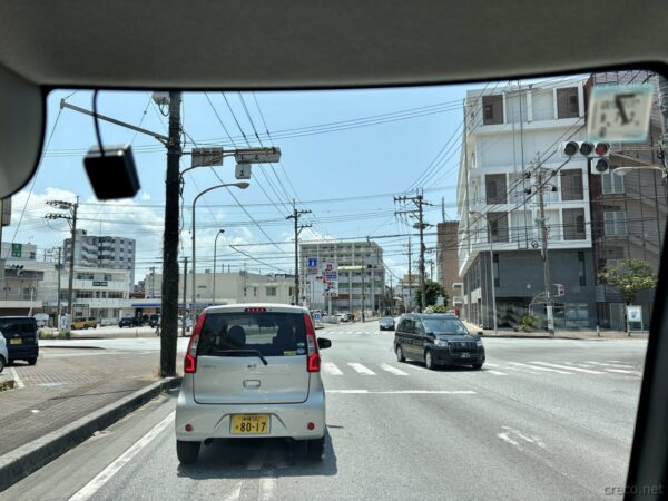 名護市街地の交差点をドライブ中の車窓。前方のダイハツミライースと街路、58号線沿いの沖縄中北部の街並み。