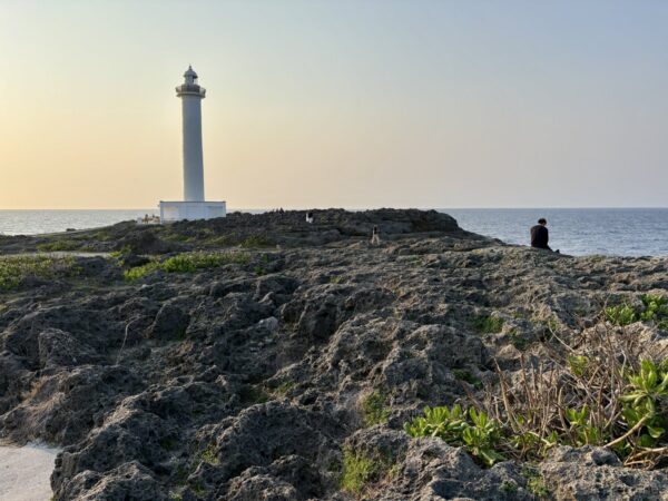 残波岬灯台を岩場から見上げる。琉球石灰岩の海岸段丘の上に立つ白亜の灯台と夕方の海、沖縄西海岸の景勝地。