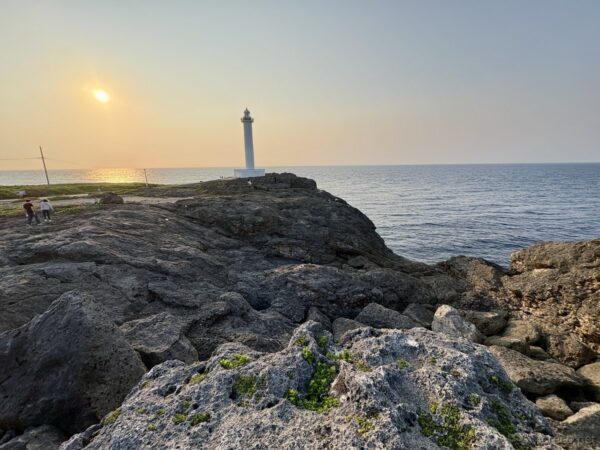 残波岬の夕日に染まる海。琉球石灰岩の岩場と低い雑草、水平線を彩るオレンジ色の空と灯台。