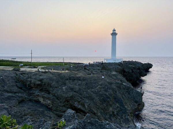 残波岬灯台と崖下の観光客たち。夕景の灯台前に佇む人影、琉球石灰岩の崖と平原のパノラマ。