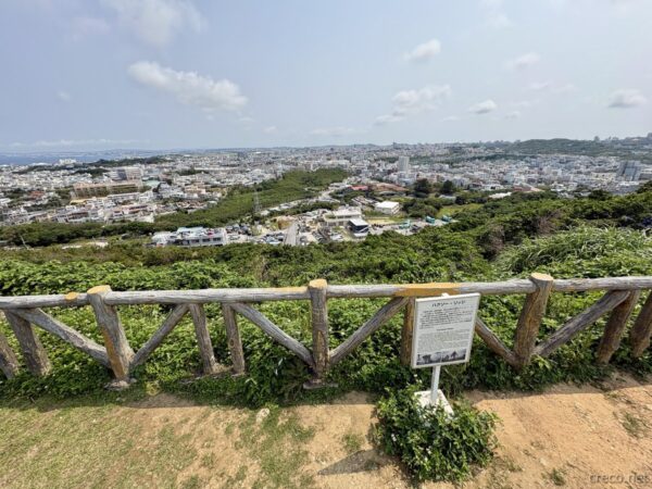 浦添城跡展望所から那覇と海を望む。木製手すりと解説板、高台から見下ろす南部の都市と海岸線のパノラマ。