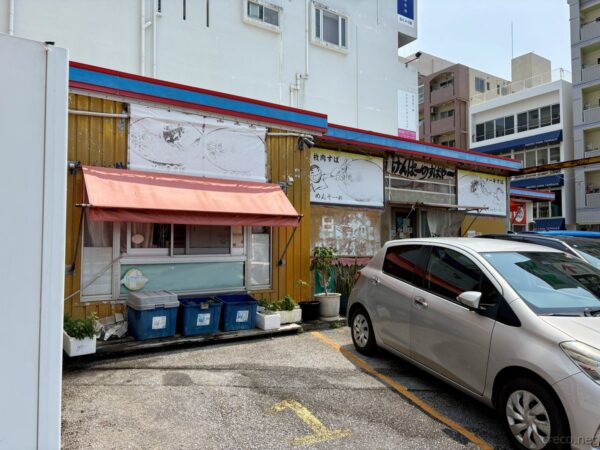 那覇市内の沖縄そば屋の外観。黄色い壁の庶民的な食堂、店先に停まる白い軽自動車、地元民に愛される沖縄そば店。