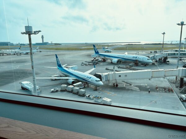 那覇空港の駐機場と全日空（ANA）機。ボーディングブリッジと747サポート車両、エプロンに並ぶブルーと白のANA機の窓景色。