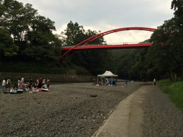 飯能河原から見上げる赤いアーチの割岩橋、河原ではBBQを楽しむ人々