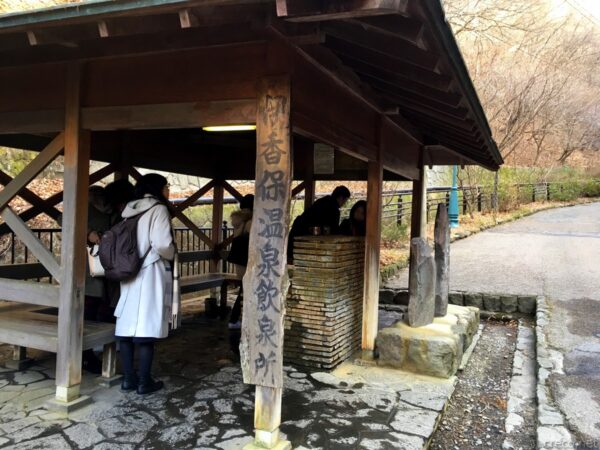 伊香保温泉飲泉所、木造の東屋風建屋に「伊香保温泉飲泉所」の墨書き看板、レンガ積みの飲泉口で観光客が試飲