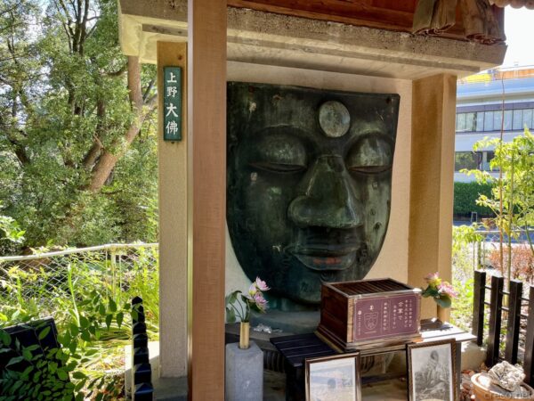 上野公園の上野大佛、緑青を帯びた青銅製の顔面レリーフが祠に祀られ、左柱に「上野大佛」の木札、足元に蓮の花