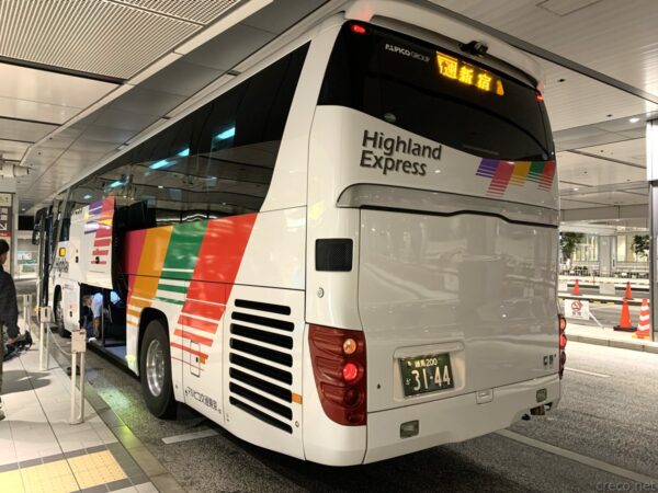 東京、屋内バスターミナルに停車する白い高速バス「Highland Express」を後方から撮影。車体側面に赤緑青などのカラフルな帯模様、リアに黄色ナンバープレート、奥に赤いコーンと通路が見える。