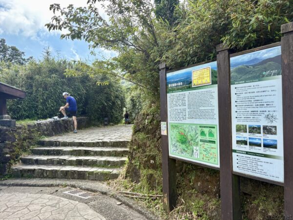 陽明山、七星山登山口(小油坑端)の入口付近。石段とともに登山道の案内看板が立ち並び、笹や樹木に囲まれた登山道が奥へと続く。青空の下で登山者が身支度している。