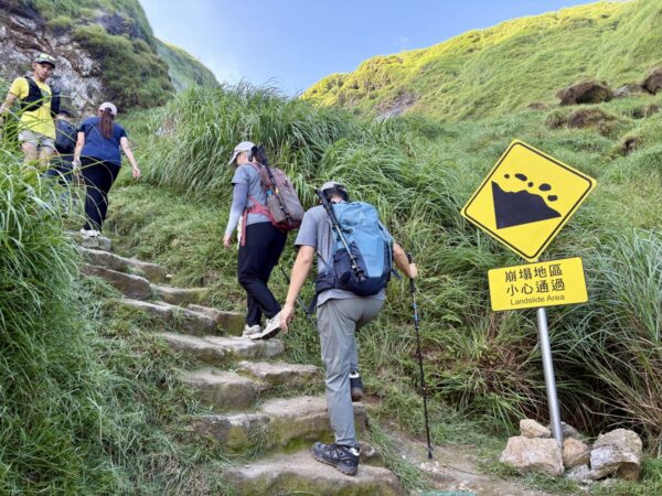 七星山の草地の急な石段を登る登山者たち。右手に落石注意「崩塌地區小心通過 Landslide Area」の黄色い警告標識が立つ。