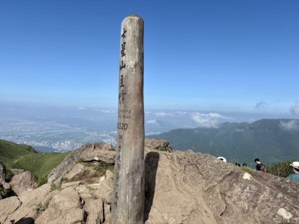 七星山主峰(標高1120m)山頂の木製標柱。青空の下、岩がちの山頂に立ち、背後に台北市街と雲海、山並みが広がる。