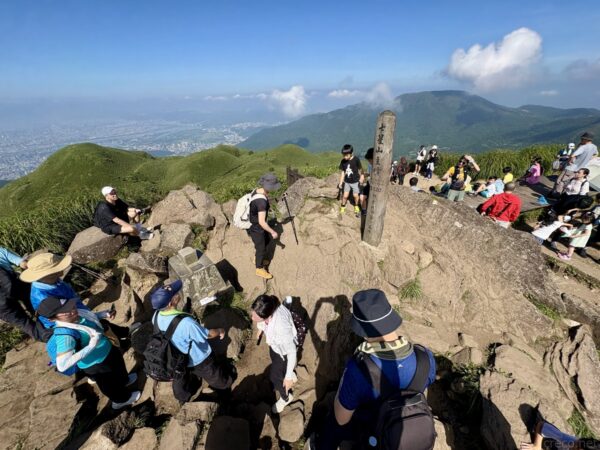 七星山山頂の岩場に腰掛け休む登山者たち。木製標柱を中心に、背後には緑の火山峰と青空、白い雲が広がる賑わいの光景。