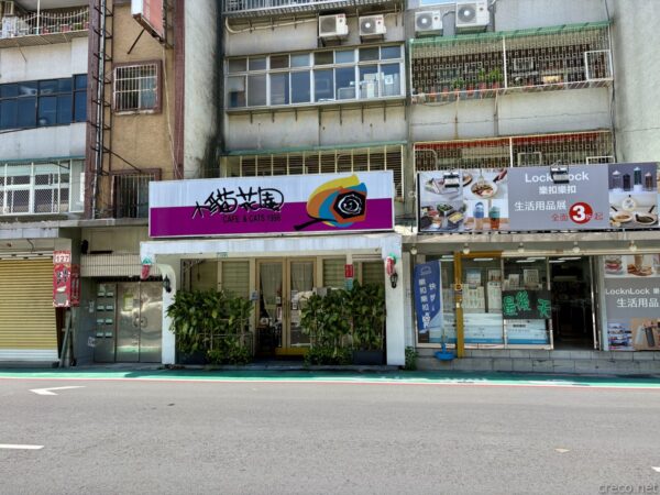 台北・芝山の猫カフェ「小貓花園」の外観。古い雑居ビル一階に紫色の看板を掲げた店構えで、観葉植物の並ぶ入口が通りに面する。隣には生活雑貨店も並ぶ。