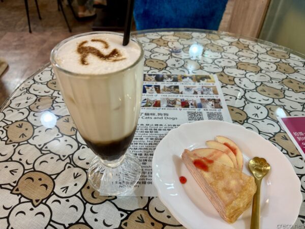 猫カフェ「小貓花園」で味わったカフェ・マキアートとケーキ。猫柄のテーブルに、猫の模様をあしらったカフェ・マキアートのグラスと、リンゴを添えたパイ風スイーツが並ぶ。