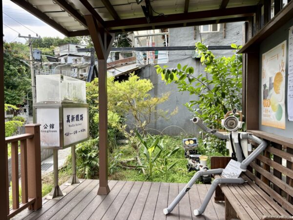 寶藏巖入ったところのウッドデッキ。手すり越しに集落と「萬新鐵路」「公館」の解説看板が見え、パイプ材で作ったロボットのアート作品が座る。