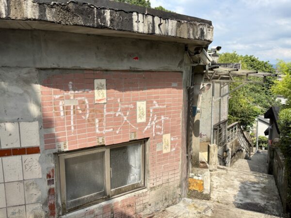寶藏巖の斜面に建つ古い煉瓦とタイルの家の裏手。かすれた文字の壁面と格子窓があり、細い路地が坂下へ延びる古い集落の一角。