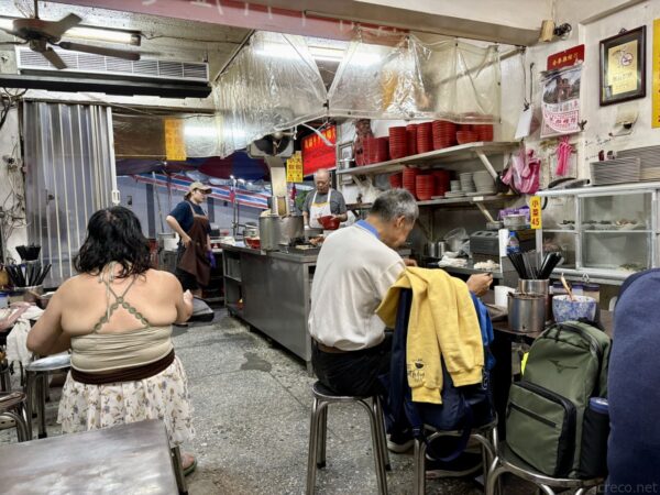 台北・城中市場の食堂「老牌牛肉拉麵大王」の店内。奥の厨房で店主が調理し、カウンター席では客が食事をする。所狭しと器や食材が積まれた庶民的な佇まい。