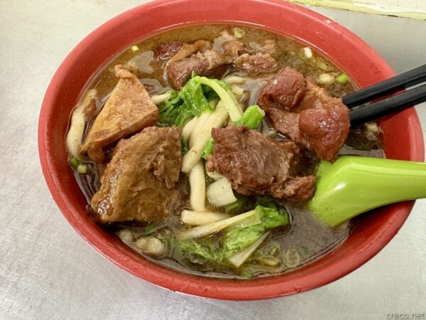 台北・城中市場「老牌牛肉拉麵大王」の牛肉麺(牛肉ラーメン)。赤い丼に牛肉や厚揚げ、青菜、麺が入り、緑のレンゲと箸が添えられた具だくさんの一杯。