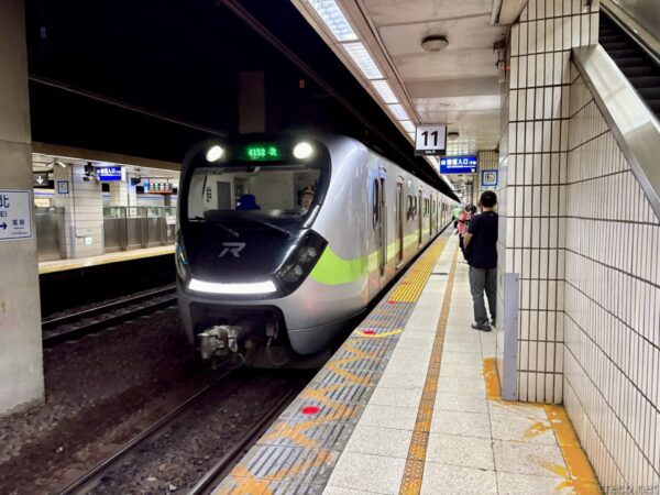 地下ホームに入線する銀色に緑帯の臺鐵通勤電車。前面に行先表示が光り、白タイルの壁際で乗客が一人待っている。11番の柱番号が見える。