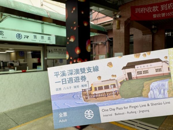 手に持った「平溪深澳雙支線一日週遊券」の一日乗車券。天燈やレトロ列車のイラスト入りで、背後に緑色の駅舎の売票処が写る。