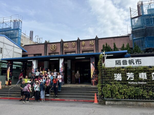 瑞芳車站の駅舎正面。「瑞芳車站 Ruifang Station」の看板を掲げた赤茶色の建物前の石段に、帽子やリュック姿の団体客が集まっている。