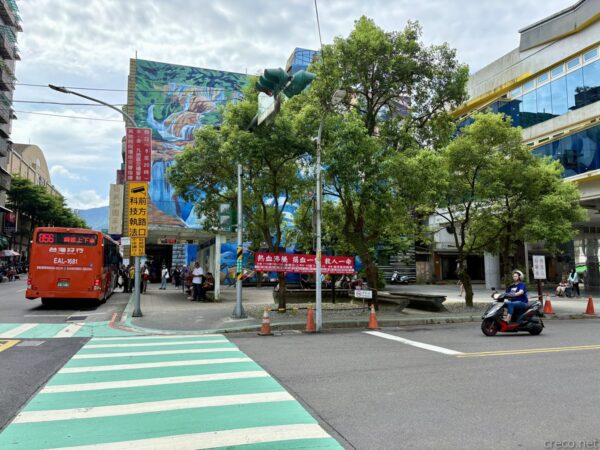 瑞芳区の区民広場付近の街角。大きな壁画が描かれたビルの前にバス停があり、路線バスやバイク、横断歩道が見える。歩道には木々が並ぶ。