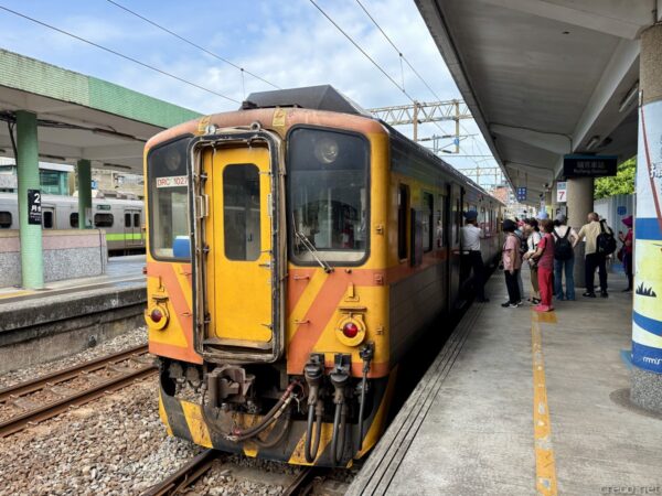 瑞芳駅ホームに停車する橙と黄色の臺鐵区間車。丸い前照灯の旧型気動車で、緑の屋根の下、ホームで乗客数人が乗車を待っている。