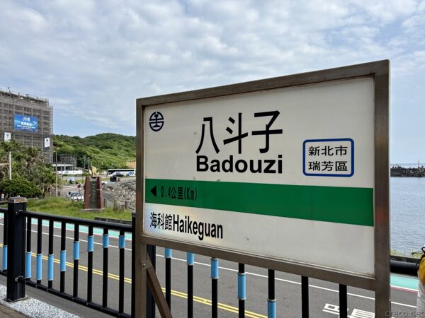 八斗子（Badouzi）駅の駅名標を大きく写した一枚。「海科館 Haikeguan」の案内と、青い柵越しに海と山が続く海辺の無人駅の雰囲気。