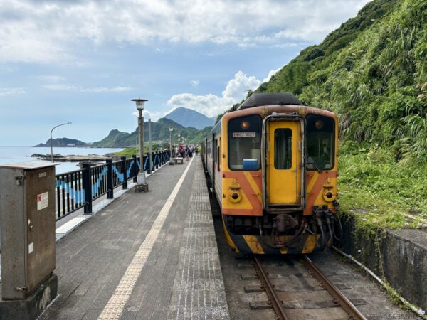 海辺のホームに停車する橙とグレーの列車の先頭部。線路が奥へ伸び、左に海と岬、右に緑の山肌が迫る八斗子駅の情景。
