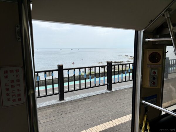 列車の開いた乗降口から見える海。ドア越しに青と黒の柵、その先に穏やかな海と防波堤、浅瀬で遊ぶ人影が小さく写る。