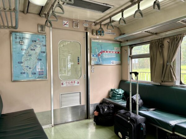 古い列車内。壁に台湾全図と路線図が掲示され、緑のクロスシートにリュックやスーツケースなどの荷物が置かれたレトロな車内。