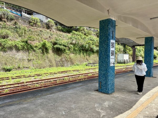 猴硐（Houtong）駅のホーム。青い花柄タイルの柱に「猴硐」の駅名と矢印。線路脇に草が茂り、白い上着に日除け帽の女性が歩いている。