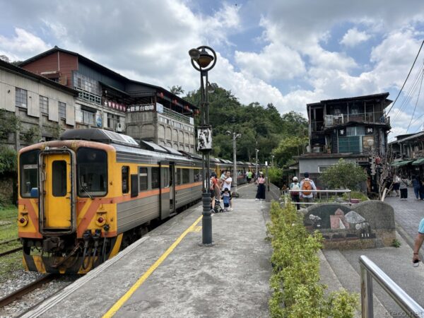 平溪線・菁桐駅のホーム。オレンジと白の旧型ディーゼル列車が停車し、街灯や乗客が並ぶ。線路脇には古い木造家屋や山の緑が見える。