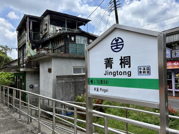 平溪線・菁桐駅の駅名標。緑と白の「菁桐 Jingtong」「平溪 Pingxi」の標識が立ち、背後には錆びたトタンや木造の古い駅舎建物と山の緑が広がる。