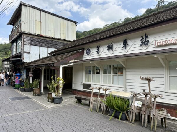 菁桐駅の駅舎正面。「菁桐車站 Jingtong」と書かれた白壁・瓦屋根の木造風駅舎の前に、竹細工のオブジェや観葉植物の鉢が並ぶ石畳の通り。左手に古い木造の物見櫓状の建物。