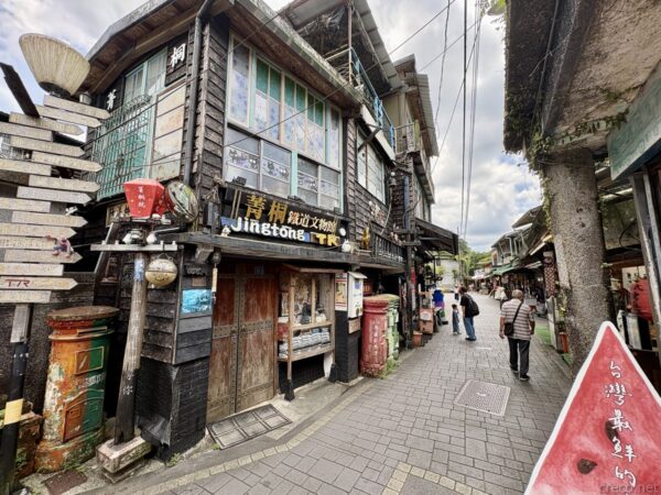 菁桐老街の商店街。「菁桐 Jingtong 鐵道文物」の看板を掲げた木造の古い店舗が軒を連ね、赤い郵便ポストや吊り飾りが並ぶ。石畳の路地を観光客が歩く。