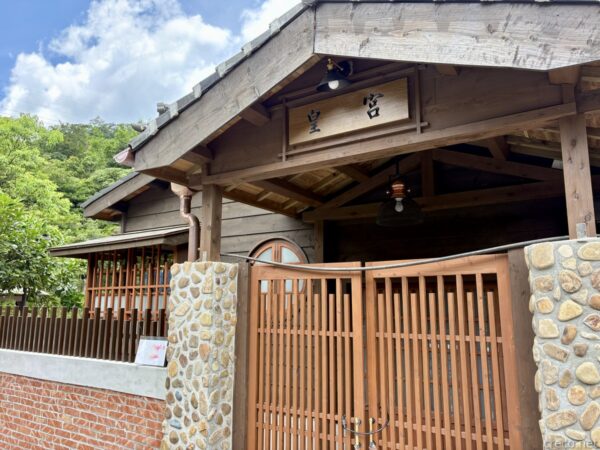 「皇宮」と扁額を掲げた木造家屋の門構え。切妻の木造庇の下に照明が下がり、石を積んだ塀と木格子の門扉が続く。背後は山の緑と青空。