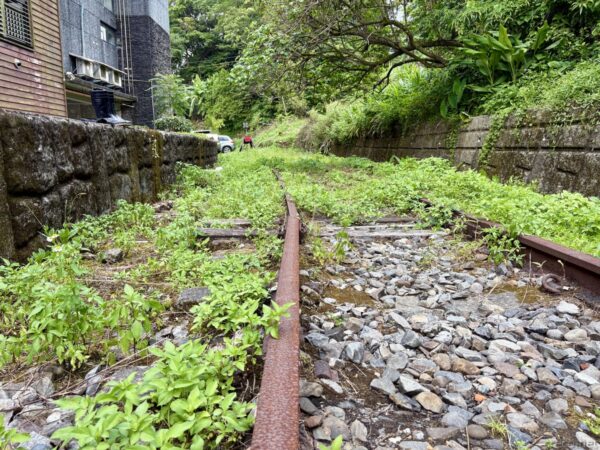 菁桐駅の線路の終点。石垣に挟まれた行き止まりの区間で、錆びたレールが雑草に覆われ、砂利とともに緑に埋もれている。左手に建物、奥に車。