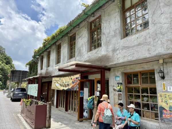 「菁桐礦業生活館」の外観。モルタル塗りの二階建て建物の軒に暖簾が下がり、緑の植栽が屋根に茂る。前を観光客が歩く。木造ではなくコンクリート造の建物。