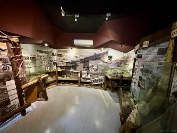 菁桐礦業生活館の展示室内部。赤茶色の壁と照明の下、鉱山の歴史写真パネルやガラスケース、木製の展示台が円形に配置されている。
