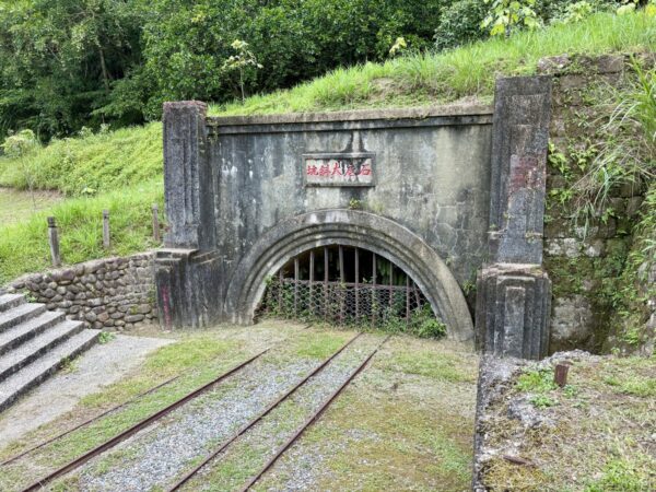 菁桐の石底大斜坑の坑口。「石底大斜坑」と赤地に記されたコンクリート造の坑門があり、格子で閉ざされたアーチ状の入口へ二本の線路が延びる。左手に石段。
