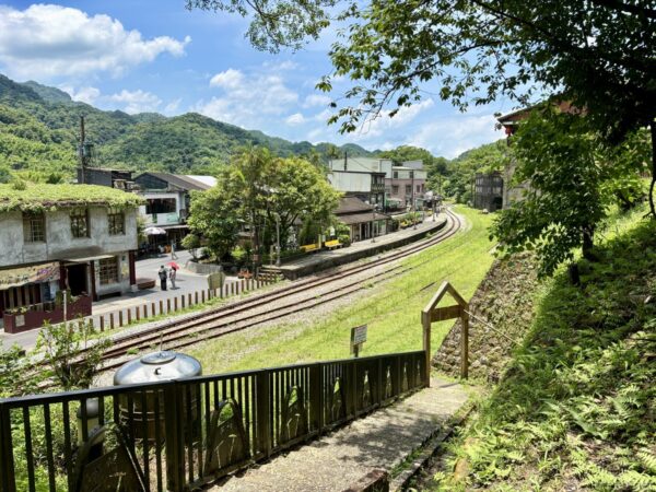 菁桐駅一帯を見下ろす眺め。緑に覆われた屋根の家屋や商店の間を線路が緩やかに曲がりながら延び、山々に囲まれた谷あいの町並みが広がる。手前に木製の欄干。