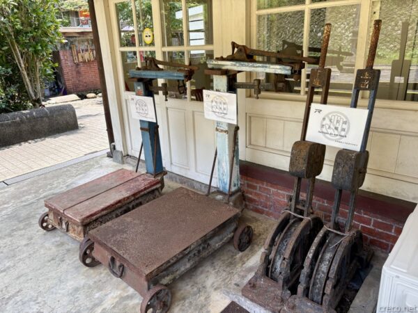 菁桐駅に展示された古い荷物用の台車と重り。車輪付きの錆びた鉄製台車や、円盤状の分銅を積んだ計量器具が、白壁・赤煉瓦の駅舎軒下に並べて置かれている。